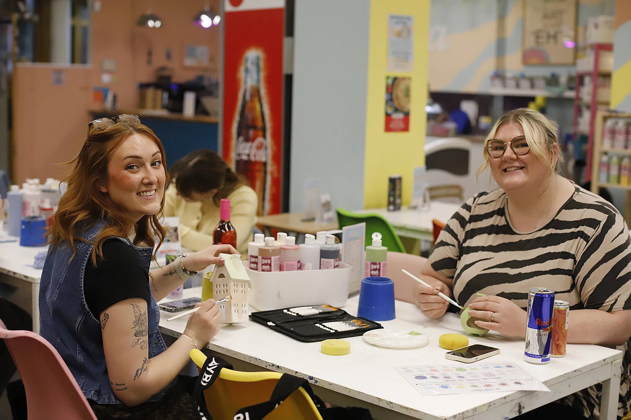 Two women smiling and painting at a white table filled with art supplies, including paintbrushes, small paint containers, and a Red Bull can, in a brightly lit indoor studio or workshop.